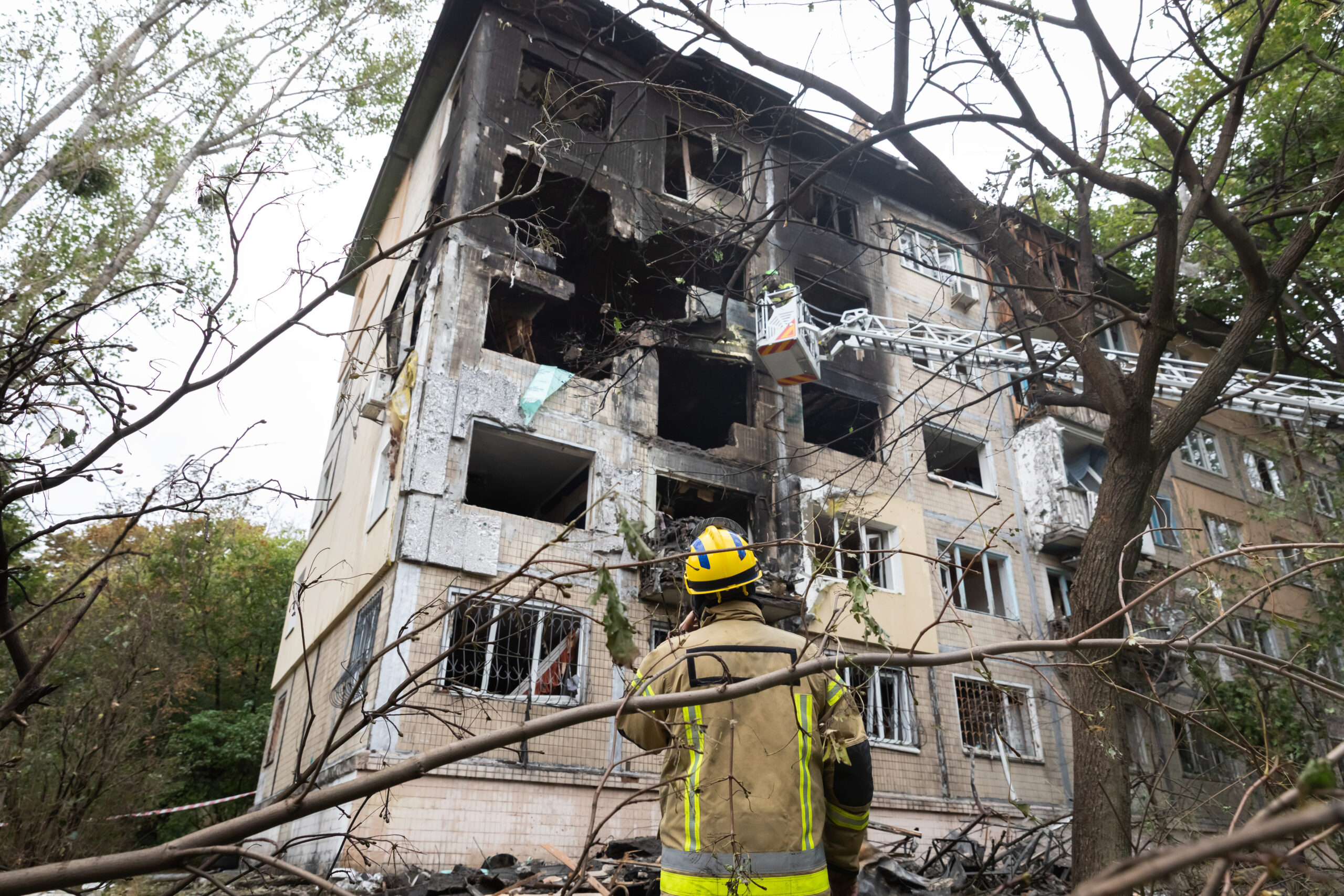 A firefighter surveying the scene of a heavily burned and structurally damaged Kyiv residential building following a Russian air attack is seen, amidst ground debris and ongoing rescue work.