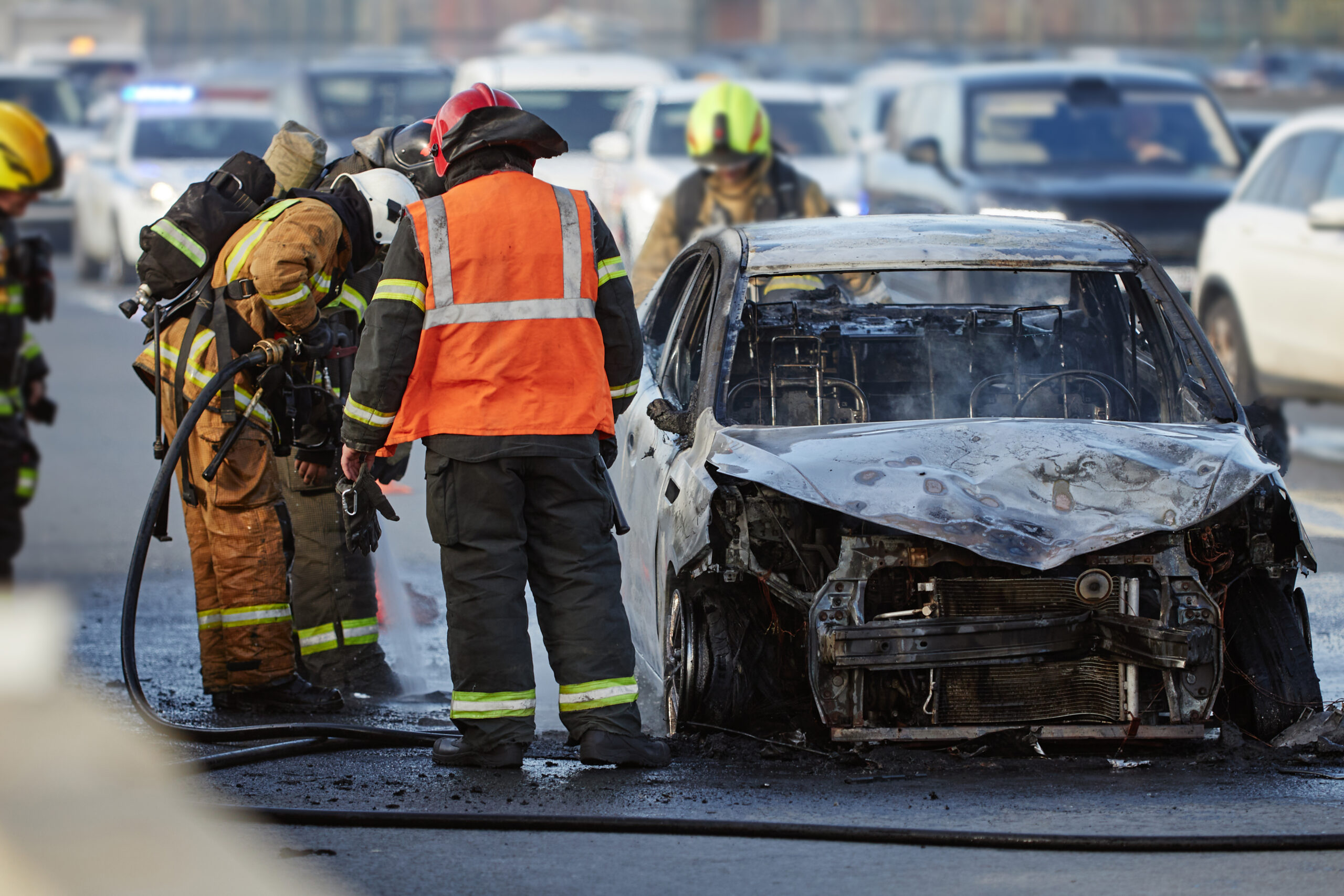 Group of firefighters and emergency responders extinguishing burning car on city street, wearing protective gear and helmets, smoke rising from damaged vehicle, traffic visible in background