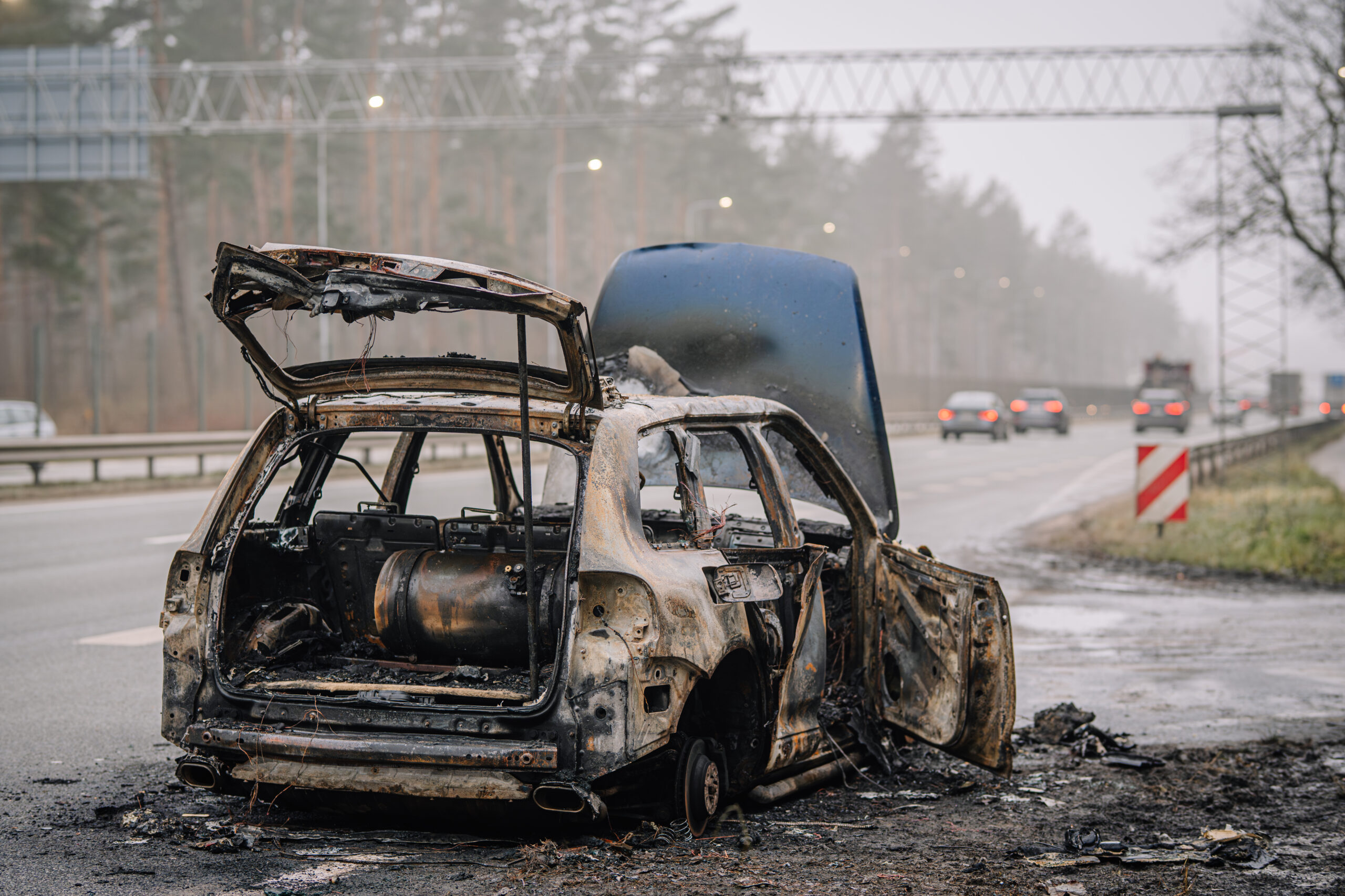 Abandoned burnt vehicle on roadside, showcasing charred remains and smoke, highlighting the consequences of fire incidents and environmental concerns